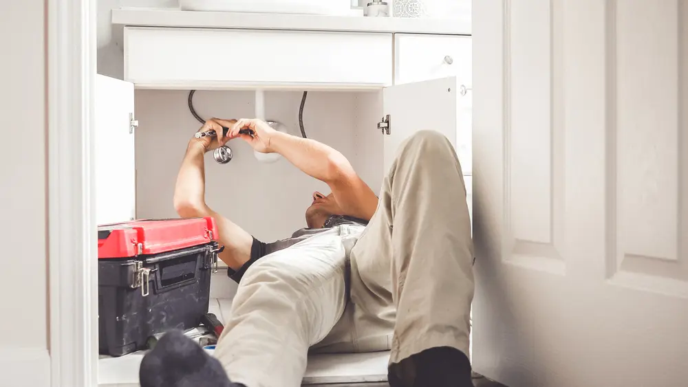 Professional plumber lying on the floor to repair a burst water pipe under a bathroom vanity sink using specialized tools.