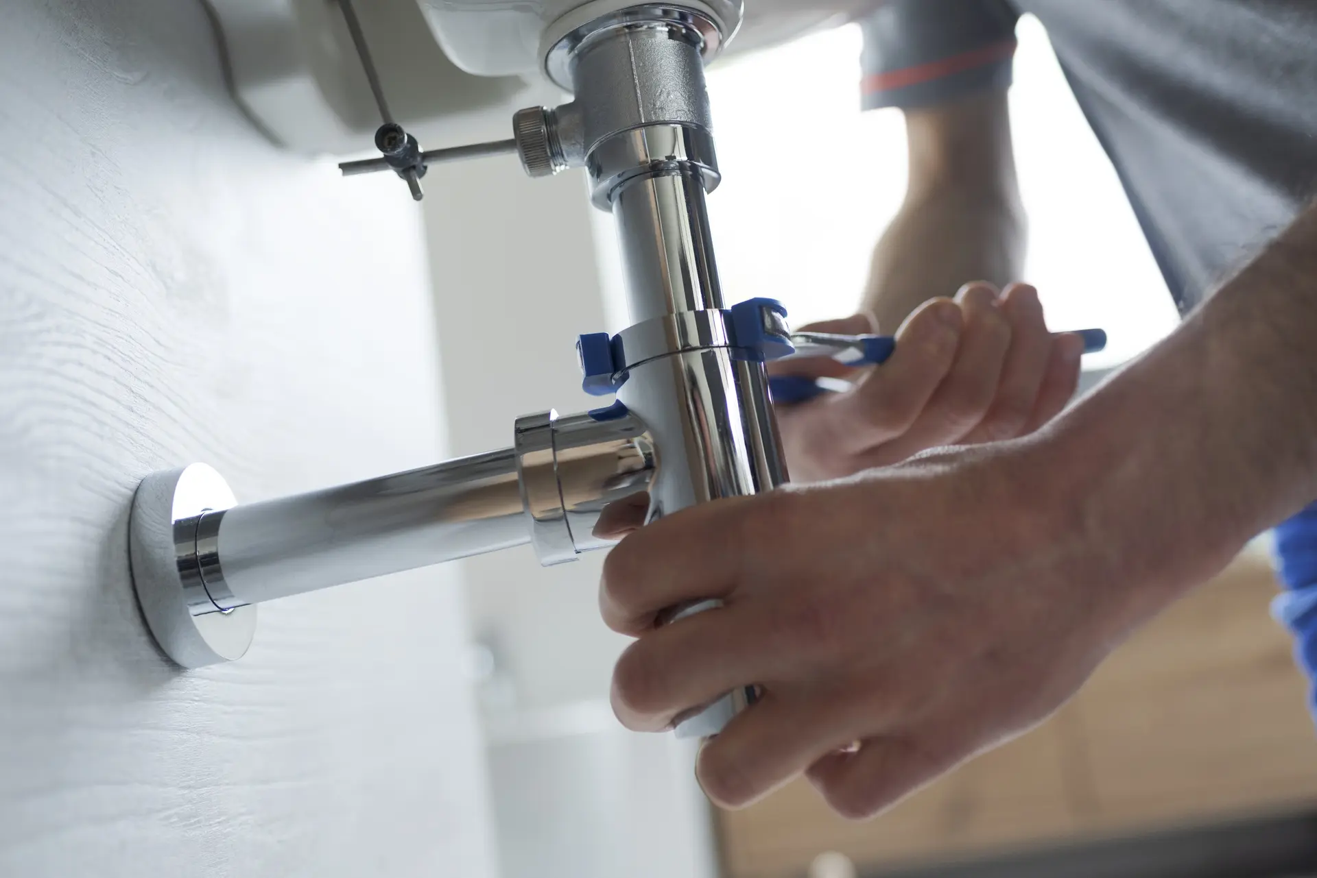 Plumber using a blue-handled wrench to tighten a chrome P-trap pipe under a bathroom sink to fix a leak