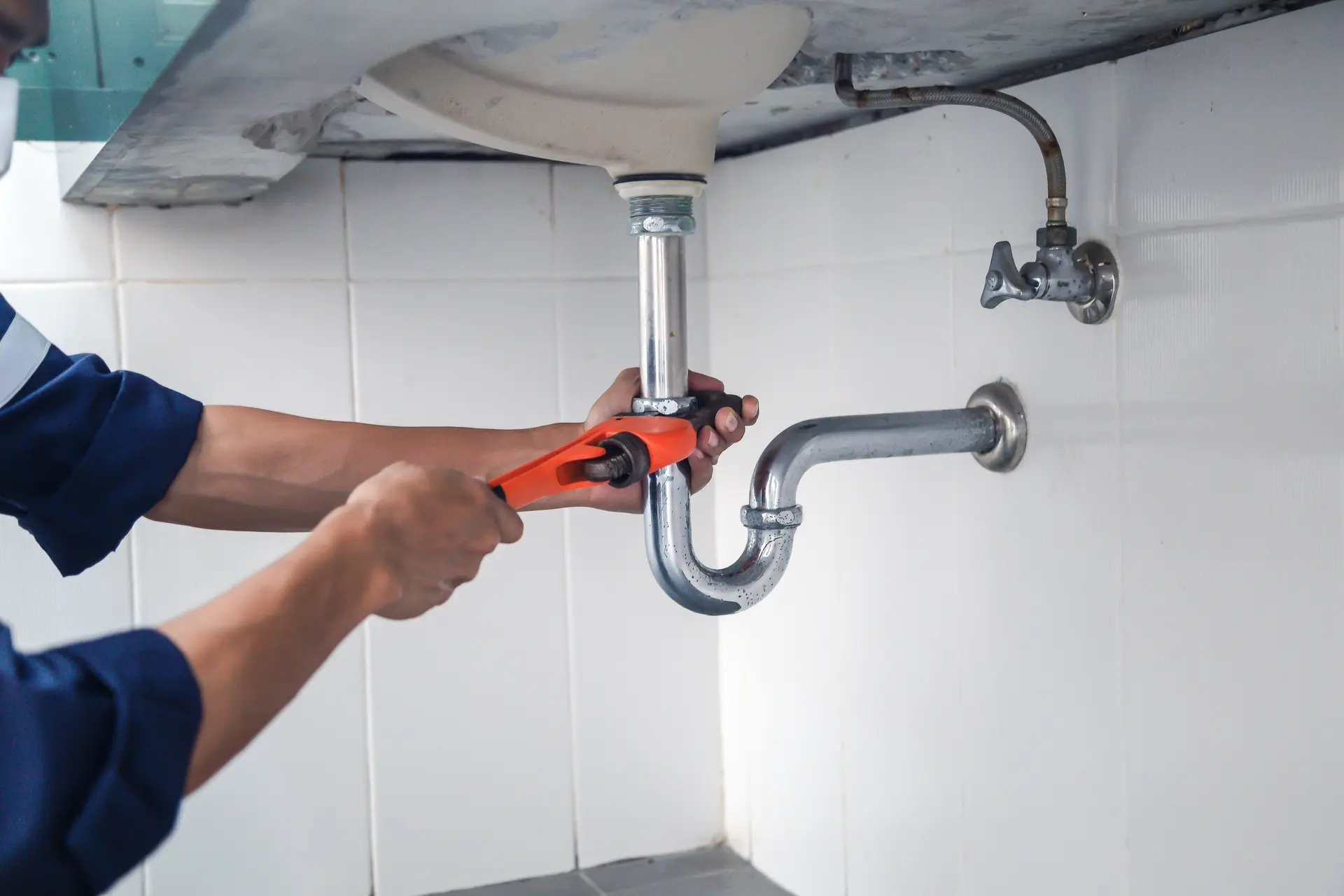 Professional plumber in a blue uniform using an orange pipe wrench to repair a chrome P-trap and metallic drain pipe under a white sink against a tiled wall.