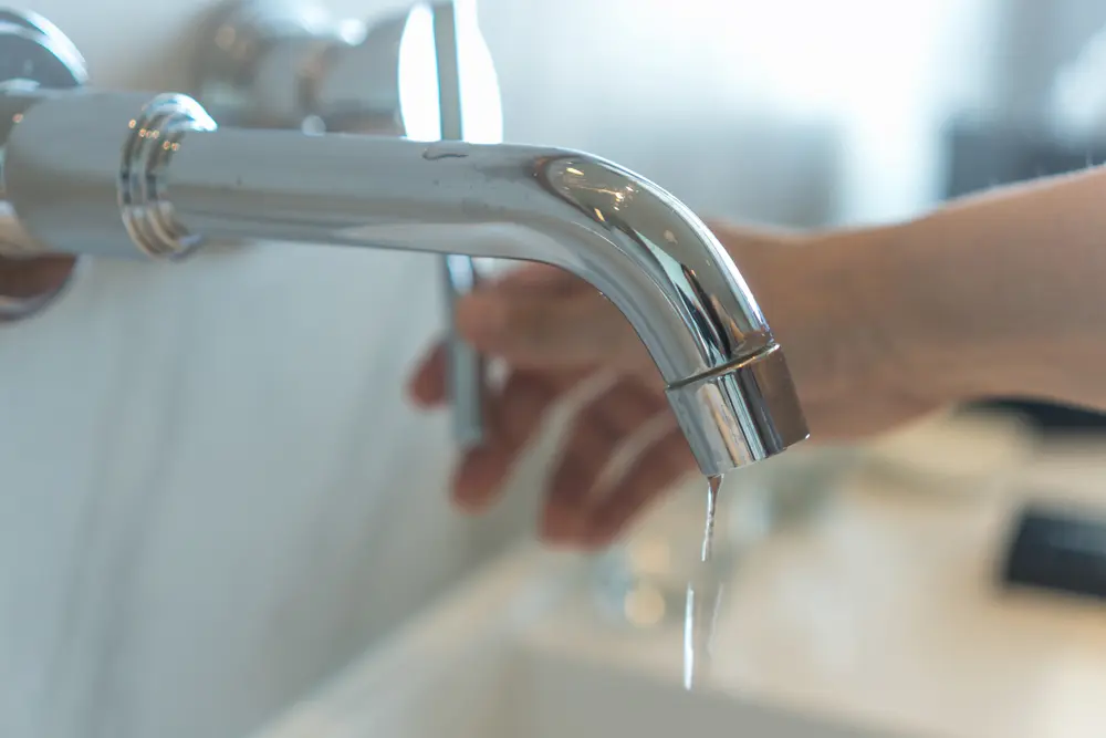 Close-up of a modern chrome bathroom faucet with a steady water drip and a person's hand in the background reaching to turn the handle.