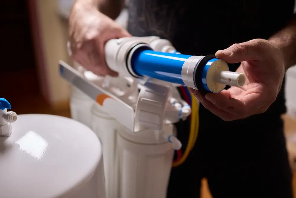 Close-up of a professional technician's hands installing a blue Reverse Osmosis membrane into a white multi-stage water filtration system housing.