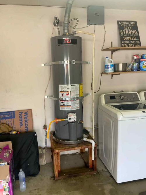 A tall grey RUUD gas water heater installed on a wooden platform in a garage, secured with metal seismic straps and a yellow gas flex line.