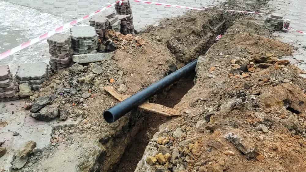 Deep trench dug through a brick-paved patio area for slab leak repair, showing a new black main water pipe installed in the ground with excavated dirt and pavers on the side.