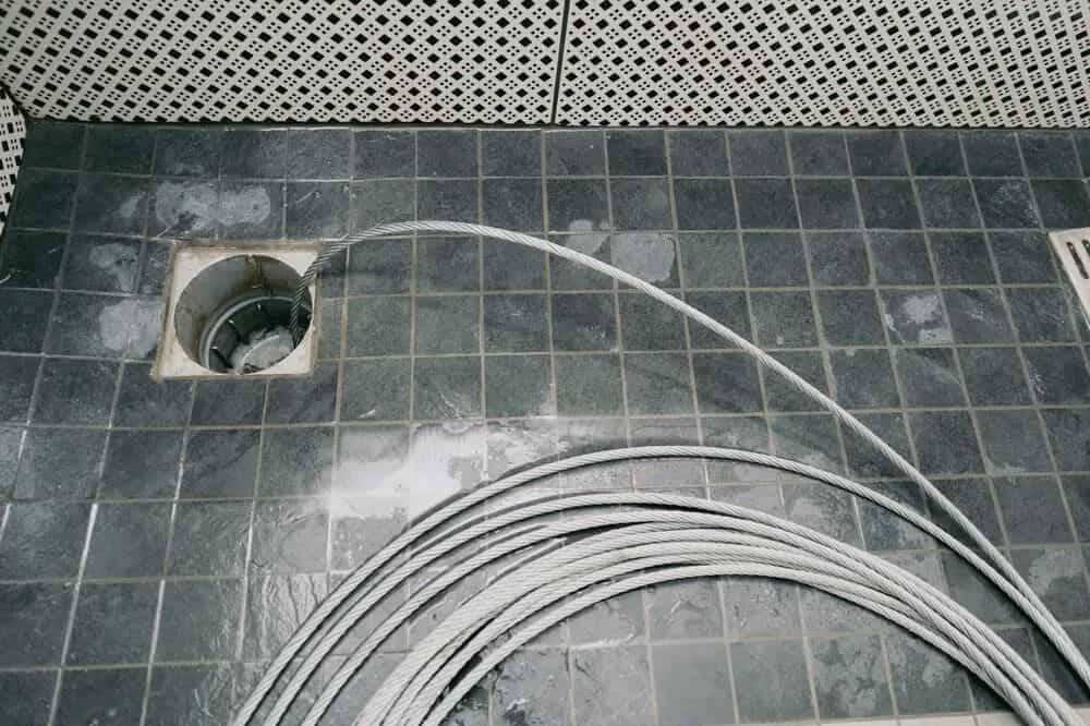 A coiled steel drain cleaning cable enters a square floor drain on dark tiled flooring during a drain clearing task. This scene represents drain and sewer services in Modesto performed by Knights Plumbing in Modesto. The metal auger cable sits across wet tiles while the tip feeds into the drain opening to clear debris inside the pipe system. Knights Plumbing provides drain and sewer services in Modesto that include floor drain clearing, sewer line cleaning, and pipe blockage removal for residential plumbing systems.