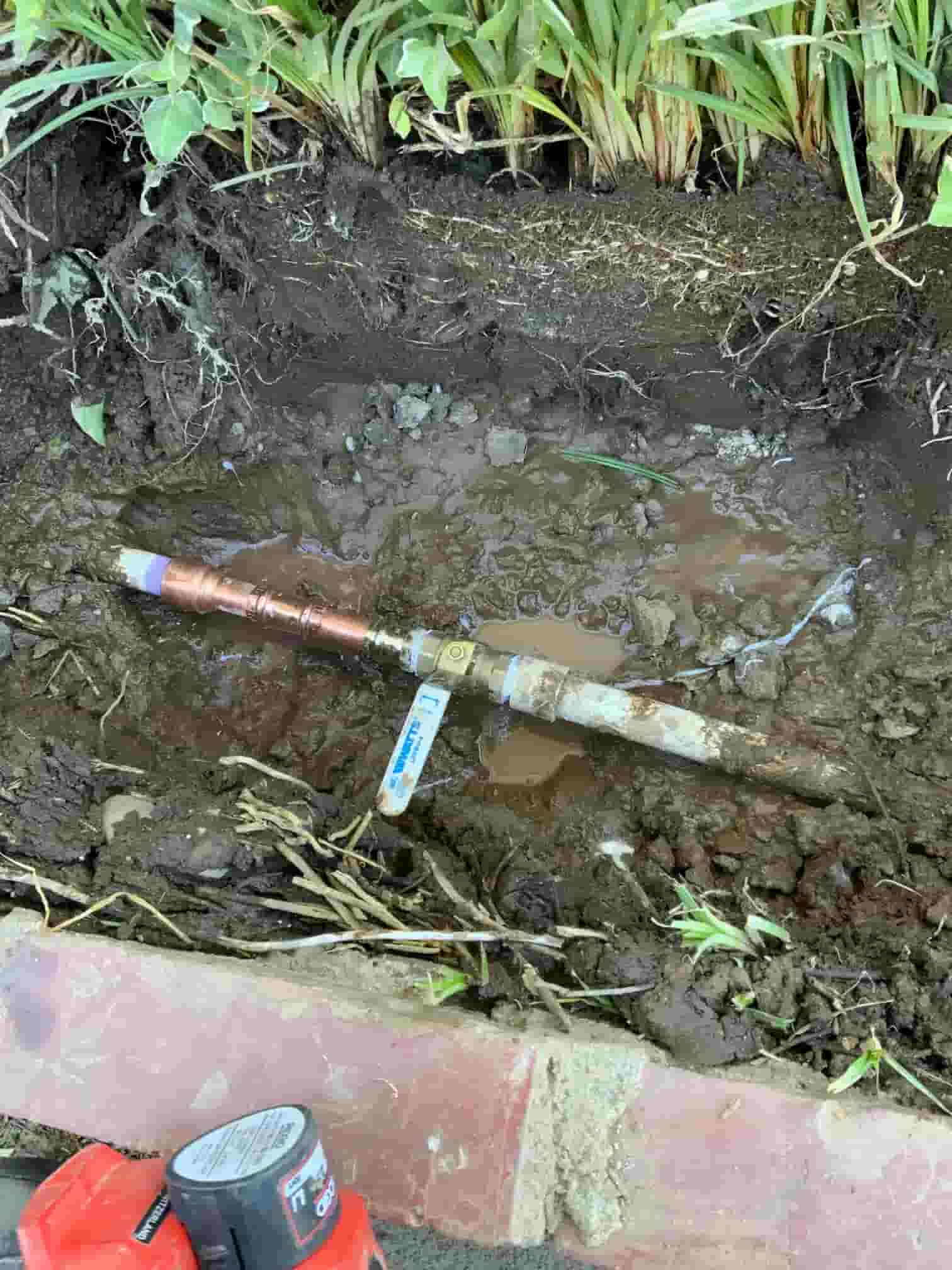 An outdoor plumbing repair in progress shows a copper pipe assembly installed in a muddy trench. The assembly includes a shut-off ball valve with a blue handle and is connected to an older pipe. The trench is dug next to a brick border and garden plants, with a red power tool partially visible in the foreground.